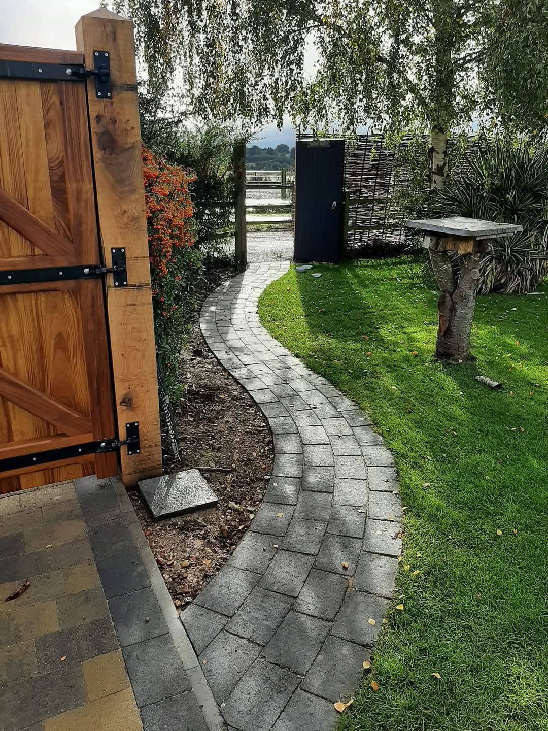 Curved block paving path beside lawn with wooden gate and weeping birch